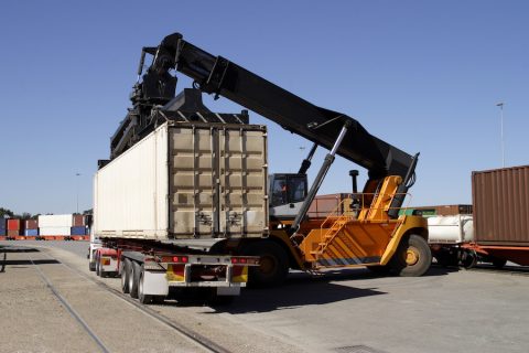 Transferring freight containers from train to truck at a city intermodal hub.  A modern container stacker loads a freight container onto a truck on a hardstand container terminal.  The container has come from a train to the left of the frame.  In the background are rail wagons (freight cars) loaded with a variety of containers. Logos and trademarks have been removed.